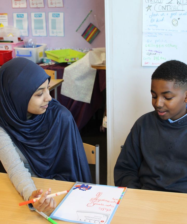 A mentor wearing a dark blue hijab and child wearing a navy hoodie engaged in conversation while completing an activity sheet in front of them